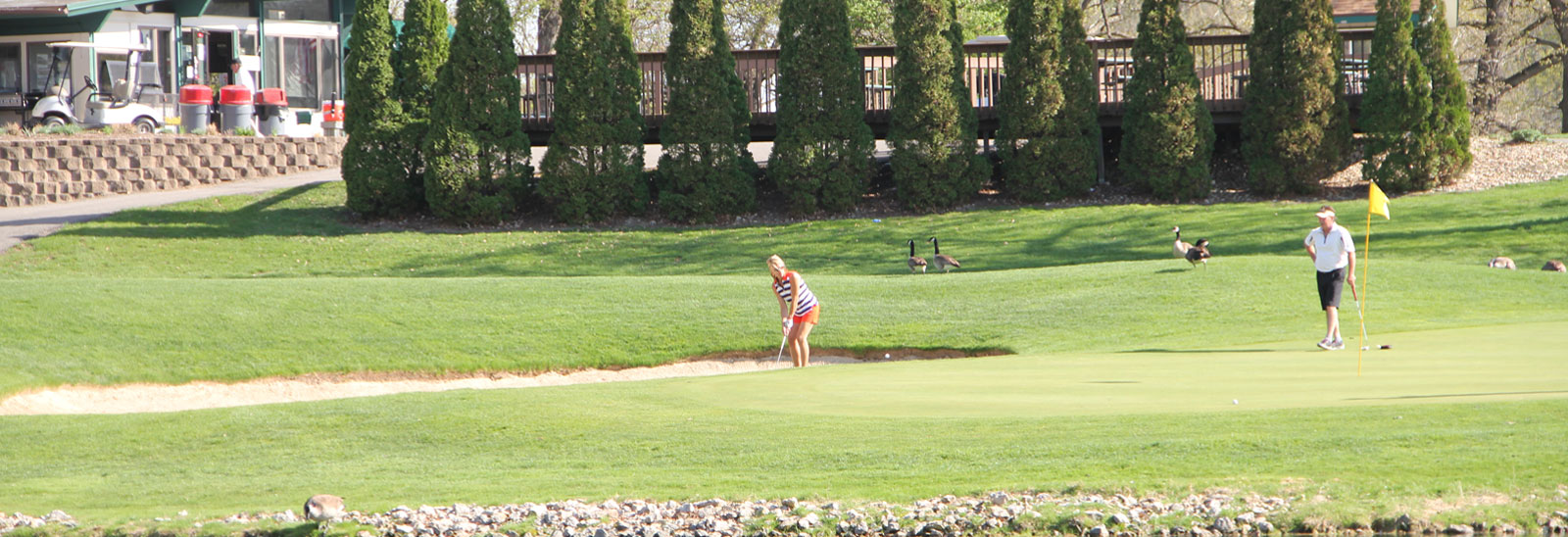 A woman chipping out of a bunker at Ellis while a man stands by the flag on the green